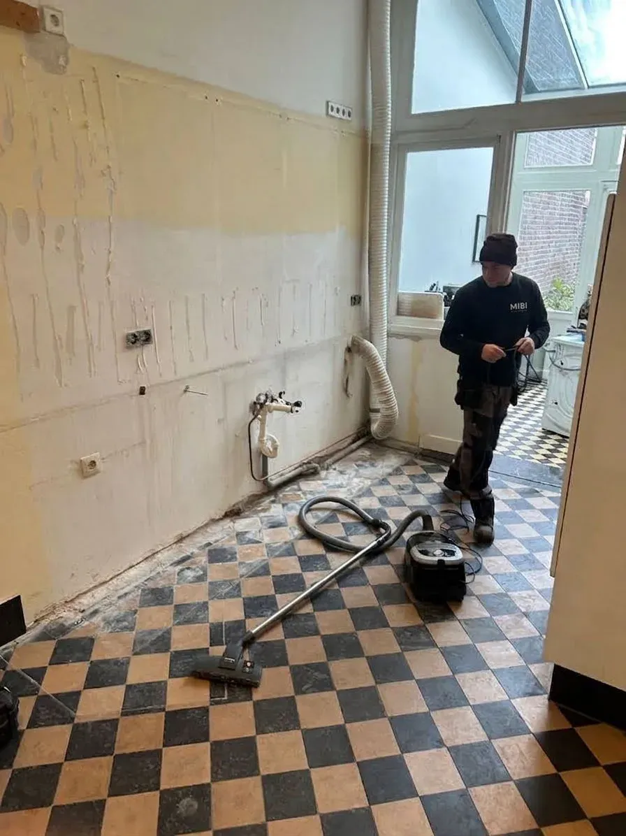 MIBI-vakman in een leeggemaakte keuken met zwart-wit tegelvloer, de stofzuiger klaar om de ruimte bezemschoon op te leveren
